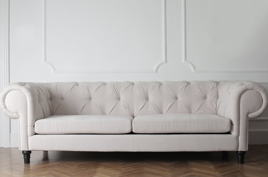 A white upholstered sofa with tufted back and rolled arms, situated in a bright, clean room with white decorative wall panels. The sofa's fabric appears spotless and well-maintained, with no visible s